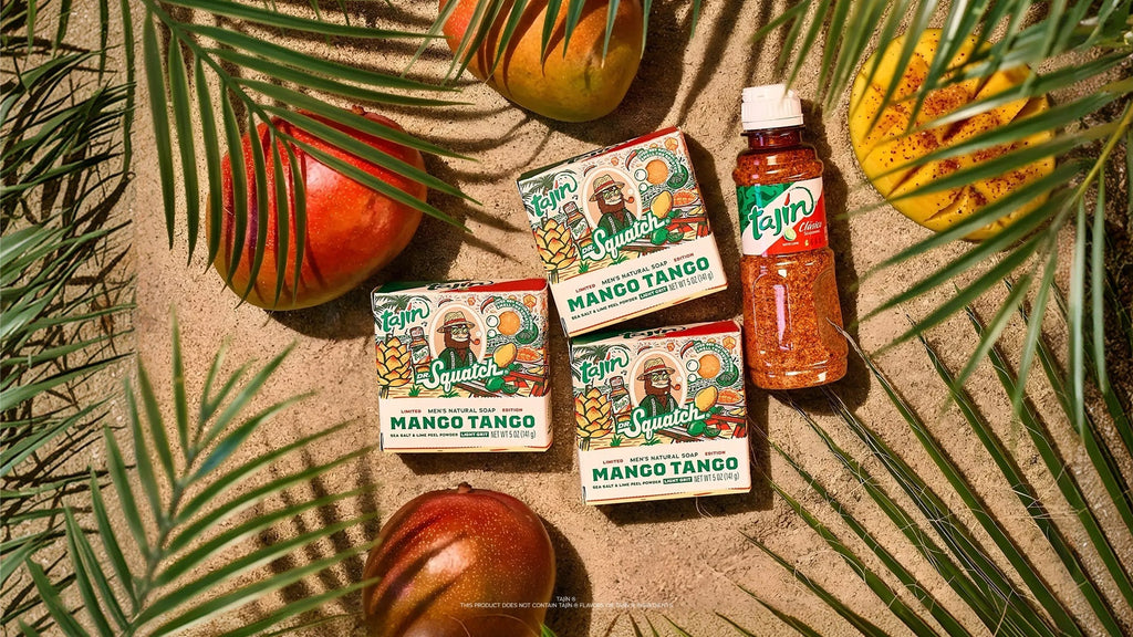 Three bars of Tajín Mango Tango soap and a bottle of Tajín seasoning arranged on sand with tropical palm leaves and mangoes.